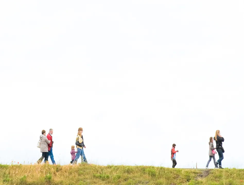 Group of people walking along a grassy path under a bright sky, including adults and children in a line across the field.