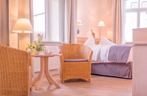 Sunlit hotel room featuring a wicker chair, round side table with a plant, and a bed with white linens and lavender bed skirt.