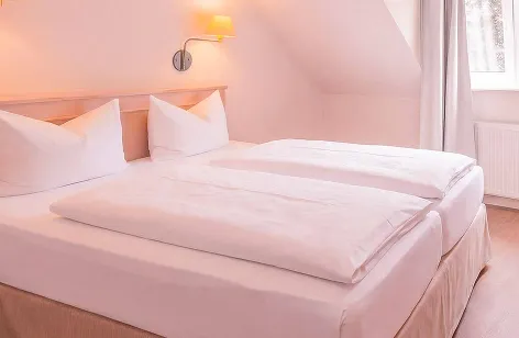 Double bed with white sheets and pillows in a bright hotel room, with a wall lamp and a window with sheer curtains nearby.