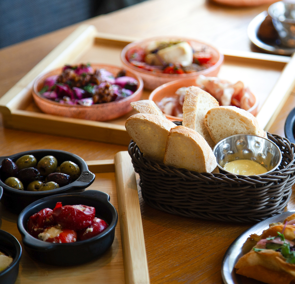 Basket of sliced baguette with a creamy dip in a metal cup, on a wicker basket, with olives and tapas bowls nearby on a wooden table