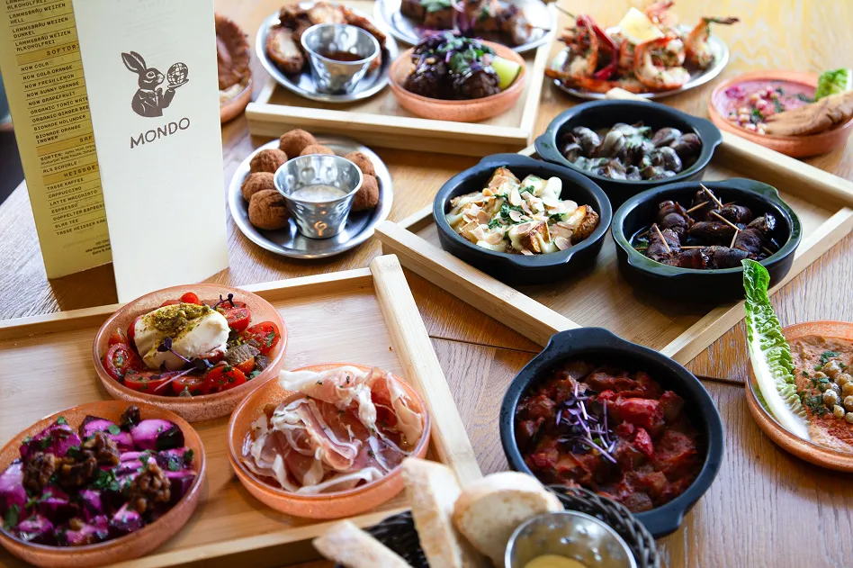 Table spread of assorted tapas: small bowls, olives, and bread baskets on a wooden table in a cozy restaurant setting